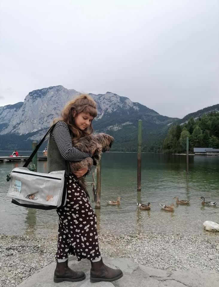 Frau mit Hund auf dem Schoss, die vor einem Bergsee steht und eine Tasche von Heidenspass um die Schulter hat
