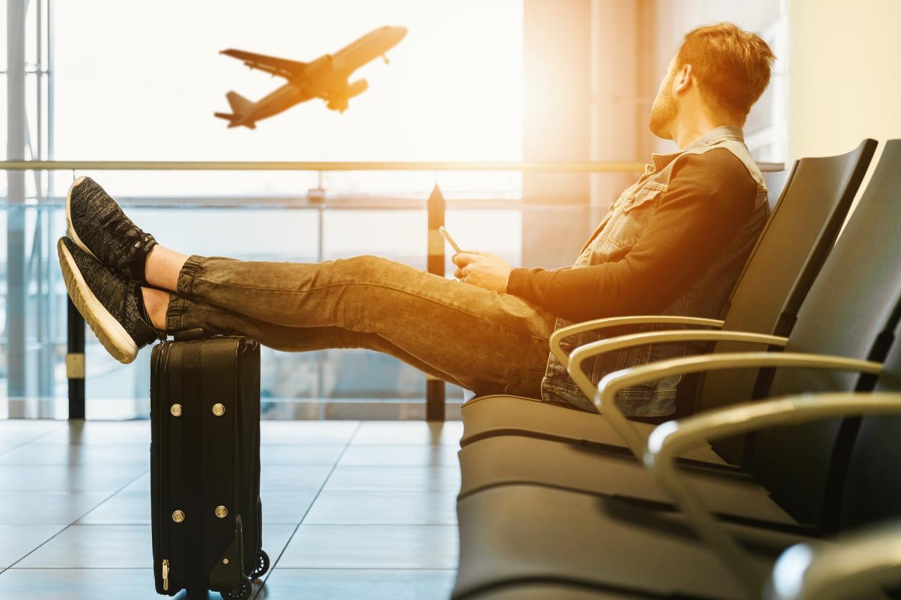 Mann, der sitzend in einer Halle am Flughafen mit seinem Koffer wartet und dabei aus dem Fenster blickt. Draußen fliegt ein Flugzeug in der Luft