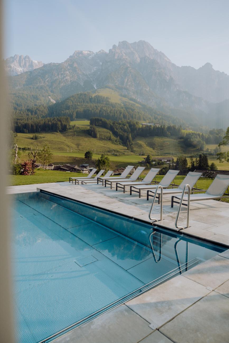 Outdoor Pool mit weißen Liegen und Blick auf grüne Hügel und Berge