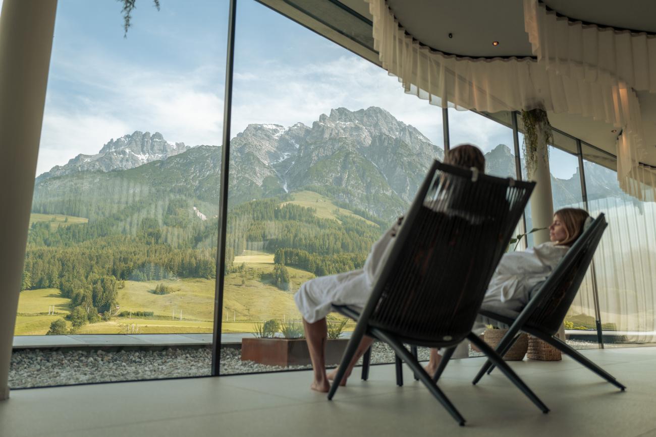 Ein und und eine Frau, die in weißen Bademäntel auf Stühlen sitzen und durch eine Glasfront auf Berge schauen.
