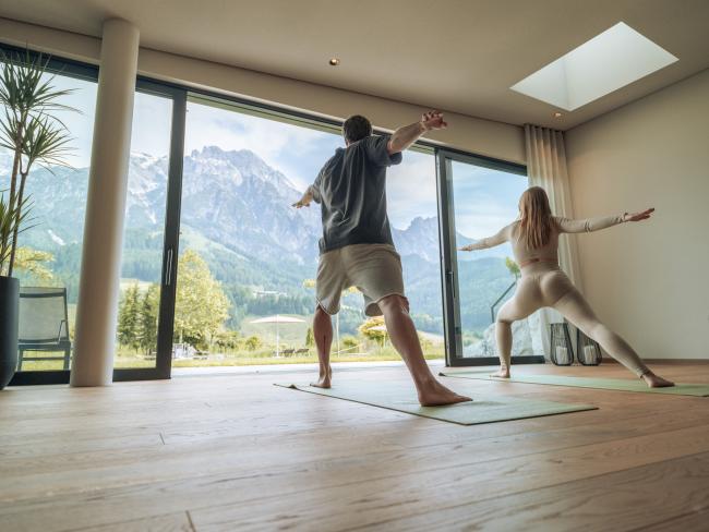 Mann und Frau die in einem offenen Raum Yoga machen und auf die Berge blicken
