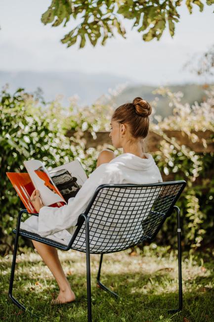 Frau, die im Bademantel auf einem schwarzen Stuhl im Garten sitzt und ein Magazin liest