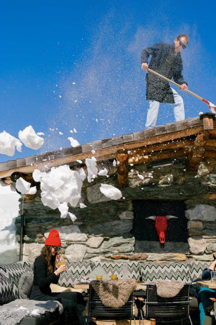 Frau genießt Drink auf Terrasse einer Skihütte, Mann schaufelt Schnee am Dach